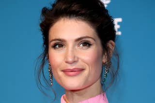 Gemma Arterton stands smiling against a blue backdrop, wearing a pink, high-neck sleeveless dress and dangling earrings