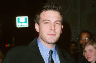 Ben Affleck, in a suit and tie, poses for a photo at a public event. Other people are visible in the background